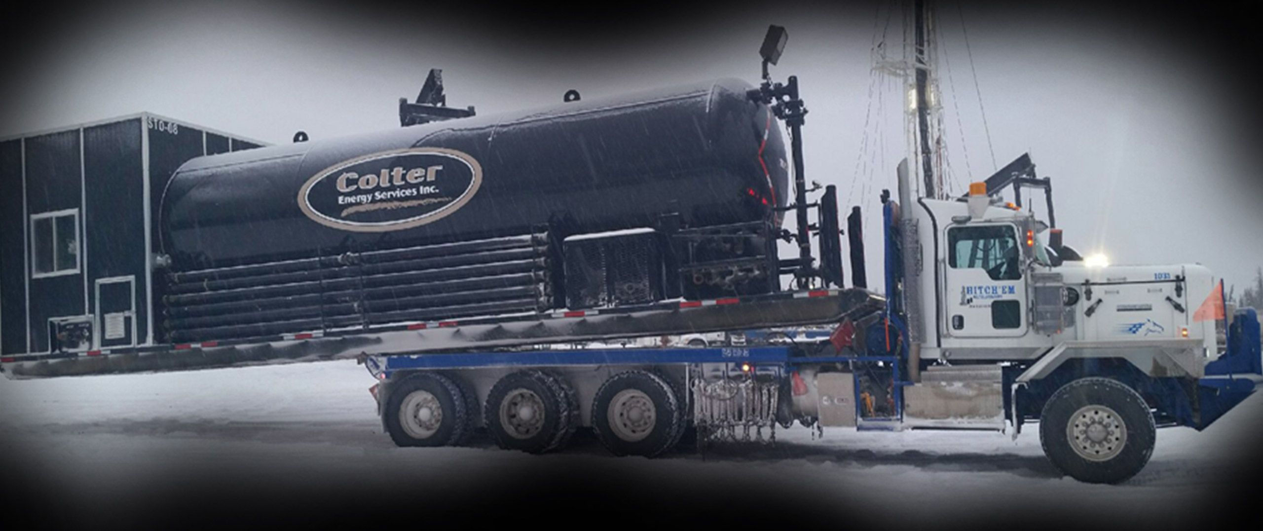A Hitch'em bed truck hauling a loaded tank across a Montney lease in winter